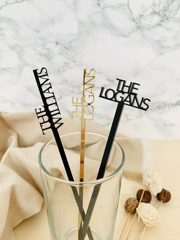 Custom acrylic drink stirrers with "THE WILLIAMS" and "THE LOGANS" in black and gold finishes, placed in a glass.