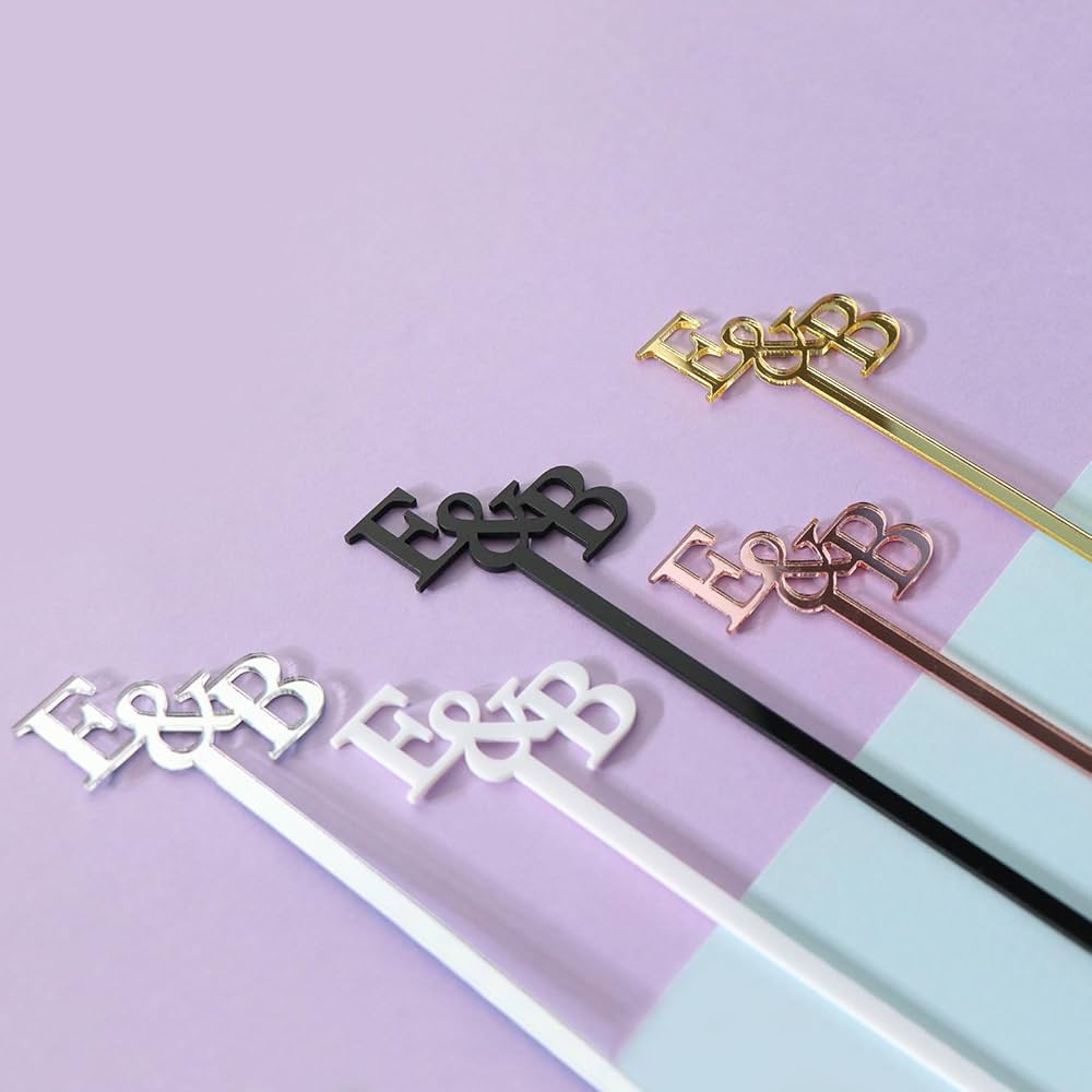 Custom acrylic drink stirrers with the initials "E & B" in multiple colors including black, white, gold, rose gold, and silver, displayed on a pastel background.